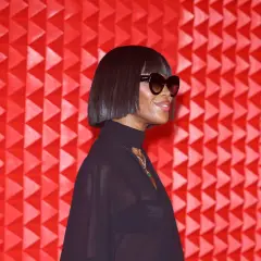 Naomi Campbell luce un corte de pelo **blunt cut** y gafas de sol, posando ante un fondo rojo texturizado.