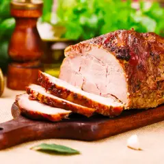 Arista di maiale morbidissima, affettata su tagliere di legno, con spezie e pomodoro sullo sfondo.