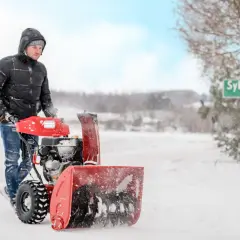 Mężczyzna w zimowej kurtce używa odśnieżarki do śniegu, by oczyścić drogę. W tle zielony znak z napisem "Syberia".