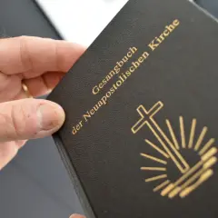 Ein Buch mit dem Titel "Gesangbuch der Neuapostolischen Kirche" wird von einer Hand gehalten. Das Symbol der Kirche, ein Kreuz mit Strahlen, ist auf dem Einband abgebildet.