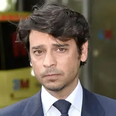 Miguel Palomo Danko, con traje oscuro y corbata, mira fijamente. Detrás, una ambulancia amarilla.
