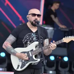 Artysta z tatuażami gra na białej gitarze, śpiewając do mikrofonu. W tle widać klawiszowca. To z pewnością niezapomniane **kombi utwory** na żywo!