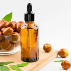 Olio di jojoba a cosa serve? Un flacone ambrato con contagocce accanto a una ciotola di frutti di jojoba e foglie verdi.