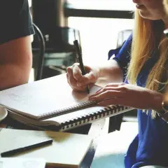Zespół pracuje nad projektem, jedna osoba pisze w notatniku, obok kubek kawy. To ich cdo stanowisko.