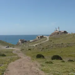 Un camino de tierra lleva al restaurante El Faro Cabo de Gata, con el mar azul y un faro en la colina.