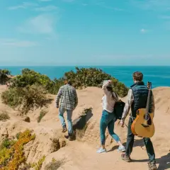 Trzy osoby wędrują ścieżką nad morzem. Jeden z nich niesie gitarę. Idealne szlaki dla dzieci na wakacyjną przygodę.