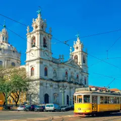 Un tranvía amarillo cruza frente a una iglesia blanca con cúpulas y torres, reflejando cómo se vive en Portugal con su encanto histórico y transporte icónico.