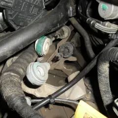 Widok na mechanizm skrzyni biegów z widocznymi linkami zmiany biegów, przewodami i elementami silnika.