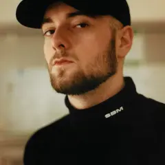 Chudy Bedoes w czarnej bluzie z golfem i czapce z daszkiem, z charakterystyczną brodą.