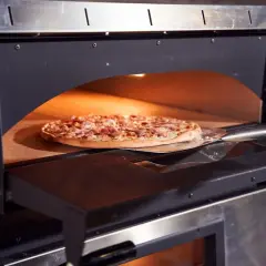 Une pizza garnie est en cours de cuisson dans un four professionnel, sur une pelle à pizza. La chaleur intense du four promet une cuisson parfaite.