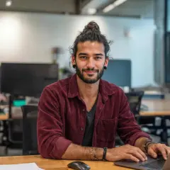 Junger Mann mit Dutt am Laptop, lächelnd. Ein zukünftiger marketing master hamburg.