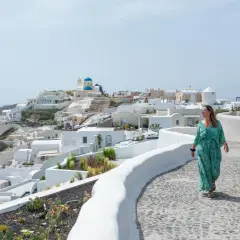 Kobieta w sukience spaceruje po Santorini, idealnym miejscu na wakacje w Grecji. Białe domy i błękitna kopuła kościoła tworzą malowniczy krajobraz.