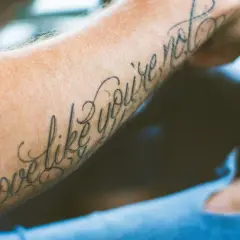 Ein Arm mit einem geschwungenen Schriften Tattoo, das die Worte "Love like you're not" trägt.
