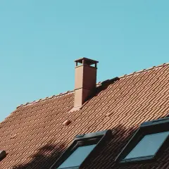 Brązowy komin na dachu z dachówki, z którego wydobywa się kondensat. Niebo błękitne.