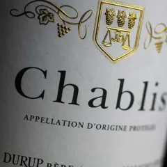 Gros plan sur une étiquette de vin Chablis, avec un blason doré orné de grappes de raisin et d'une balance. L'appellation d'origine protégée est clairement visible.