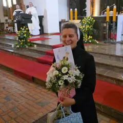 Radosna zakonnica z bukietem kwiatów i kartką z nutami. W tle msza święta, 25 października.
