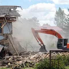 Pomarańczowy koparka prowadzi rozbiórkę budynku, wzbijając tumany kurzu. Ruiny i drewno leżą na ziemi.