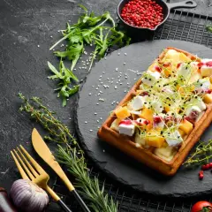 Gaufre salée garnie de fromage, de dés de mangue et de pousses, sur une ardoise noire.