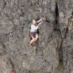 A man is climbing a steep, rocky cliff face. He's wearing shorts and a backpack, looking like he's truly "cliffed out" and facing a challenging ascent.