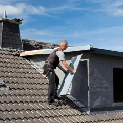 Un couvreur installe un panneau sur un toit, près d'une lucarne. Travaux d'isolation en cours.