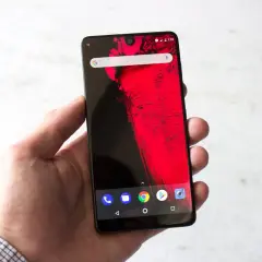 Dłoń trzyma smartfon z wycięciem (notch) na górze ekranu, wyświetlający czerwony wzór i ikonki Androida.