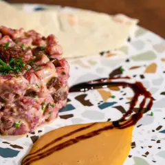 Delicioso tartar de salchichón, con trocitos de frutos secos y cebollino fresco, servido en un plato moderno con salsas.