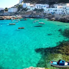 Boats in a turquoise bay near white buildings, a picturesque scene reminiscent of the charm found near international airports in Sicily.