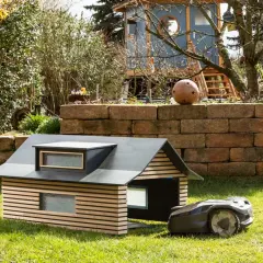 Ein moderner, selbstgebauter Rasenroboter-Unterstand mit Holzlamellen und Dachfenstern. Der Rasenroboter parkt daneben.