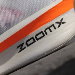 Biały but sportowy z pomarańczowym paskiem i napisem ZOOMX.