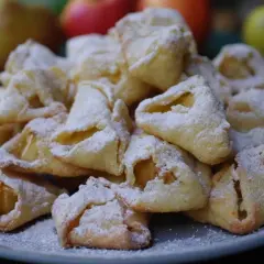Stos ciasteczek serowych z jabłkami, oprószonych cukrem pudrem, na talerzu. W tle jabłka.