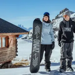 Para trzymająca deski snowboardowe przed drewnianą chatką w górach.