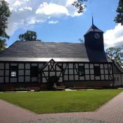 Kościół z wieżą zegarową, otoczony zielenią i ławkami. W tle niebo z chmurami. Miejsce pielgrzymek do Matki Boskiej Sianowskiej.