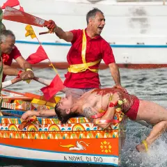 Uomo in barca siciliana, coperto di rosso, durante le feste. Un rematore con remo insanguinato.