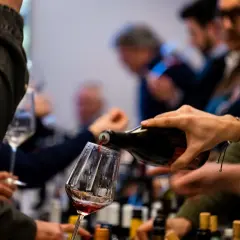 Versando vino rosso in un calice, si apprezzano le caratteristiche del vermentino. Persone in attesa, bottiglie sullo sfondo.