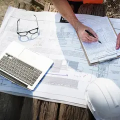 Ręce kierownika budowy nad planami, laptopem i kaskiem. Zastanawiasz się, ile kosztuje kierownik budowy?