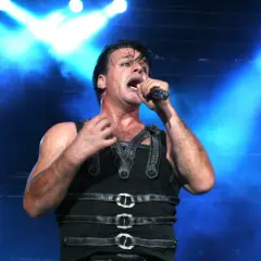Till Lindemann na koncercie, w czarnym, skórzanym stroju, śpiewa do mikrofonu na tle niebieskich świateł.