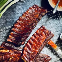 Saftige spare ribs glänzen in BBQ-Sauce auf einem Grillrost. Frühlingszwiebeln und eine Sauce-Schale deuten auf ein köstliches Mahl hin.