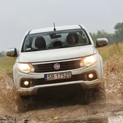 Biały Fiat Fullback z impetem wjeżdża w błotnistą kałużę, wzbijając fontanny brudu.