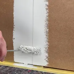 Hand streicht mit einer Farbrolle weiße Farbe auf folierte Paneele. Der Boden ist mit Malervlies und Klebeband geschützt.