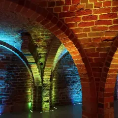 Kamienne łuki i ceglane ściany tworzą tajemniczą atmosferę. Piwnica romańska oświetlona jest kolorowymi światłami, tworząc magiczny nastrój.