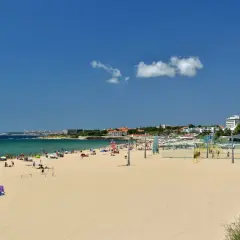 Amplia playa de arena dorada cerca de Lisboa, con gente disfrutando del sol y el mar. Hay una cancha de voleibol y edificios al fondo.