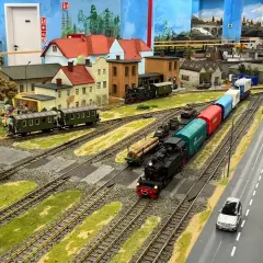 Miniaturowa kolejka z parowozem i wagonami, jak w Energylandii, przemierza malowniczy krajobraz z domkami i drogą.