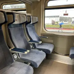 Wygodny wagon z przedziałami, z niebieskimi siedzeniami i stolikami. Za oknem deszczowa pogoda i osoba z parasolem.