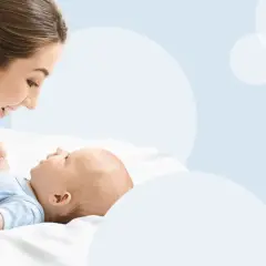 Uśmiechnięta mama pochyla się nad leżącym na plecach niemowlęciem, trzymając je za rączki.