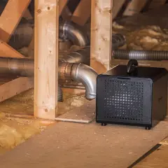 Svart avfuktare krypgrund placerad bland träbjälkar och isolering, med ventilationsrör i bakgrunden.
