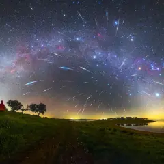 A lone figure watches a spectacular display of shoot stars streaking across the Milky Way above a tranquil lake and grassy hills.