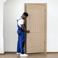 Un ouvrier en salopette bleue manipule la poignée d'une porte intérieure en bois clair, prêt à la changer.