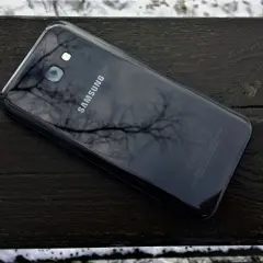 Czarny Samsung SM-A520F leży na drewnianej ławce, odbijając gałęzie drzew.