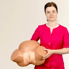 Kobieta w różowej bluzce trzyma model miednicy, prezentując anatomię do nauki masażu warg sromowych.