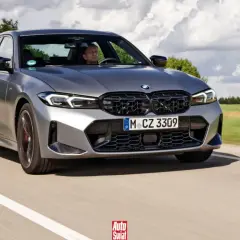Szare BMW 320d jedzie po drodze, za kierownicą siedzi mężczyzna. W tle drzewa i chmury.